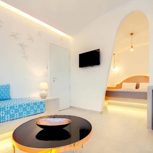 Open-plan suite interior with arched bedroom alcove, sofa and flat-screen TV at Emerald Collection Suites, Santorini